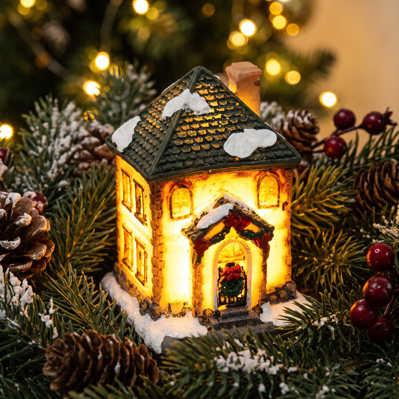 Close view of the lit facade and snow dusted roofline of a Christmas village house ornament among fir branches and pine cones