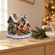 Flat acrylic Christmas snowhouse decoration on a light oak console beside fir branches