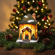 Lighted Christmas village house ornament on fir branches with a Christmas tree and golden fairy lights behind it
