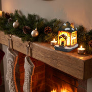 Lighted Christmas village house on a wooden mantelpiece with a fir garland, baubles, knitted stockings and a lit fire
