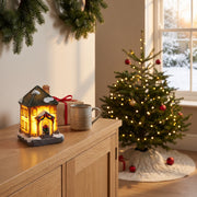 Lighted Christmas village house on a light oak sideboard in a living room dressed for Christmas, a lit tree and a snowy window behind