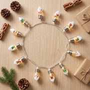 Overhead flat lay of the Christmas bulb string lights coiled on a pale oak table with pine cones and wrapped gifts