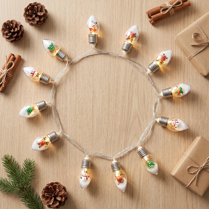 Overhead flat lay of the Christmas bulb string lights coiled on a pale oak table with pine cones and wrapped gifts