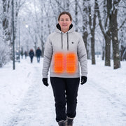 Woman walking in a snowy park wearing the gray heated hoodie, with front heating zones illustrated as a schematic red overlay, not a printed design.